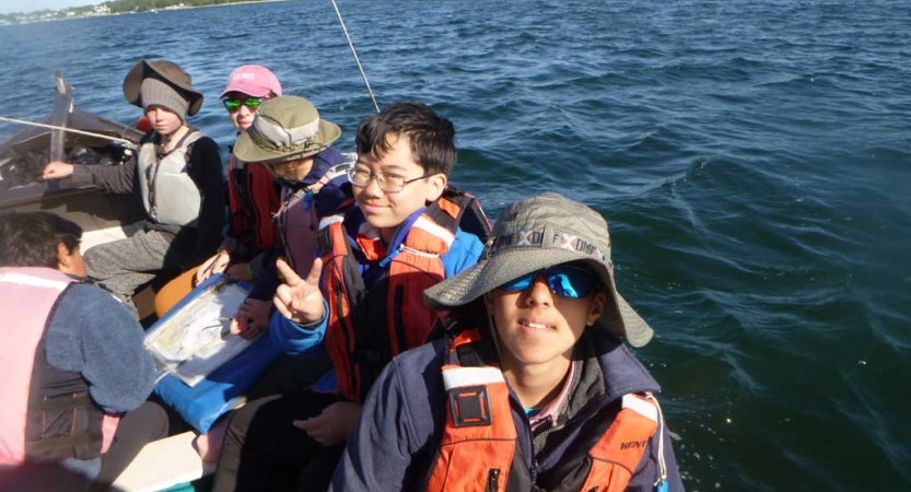 Young people wearing life jackets sit in a sail boat. Some of them are looking at the camera. 